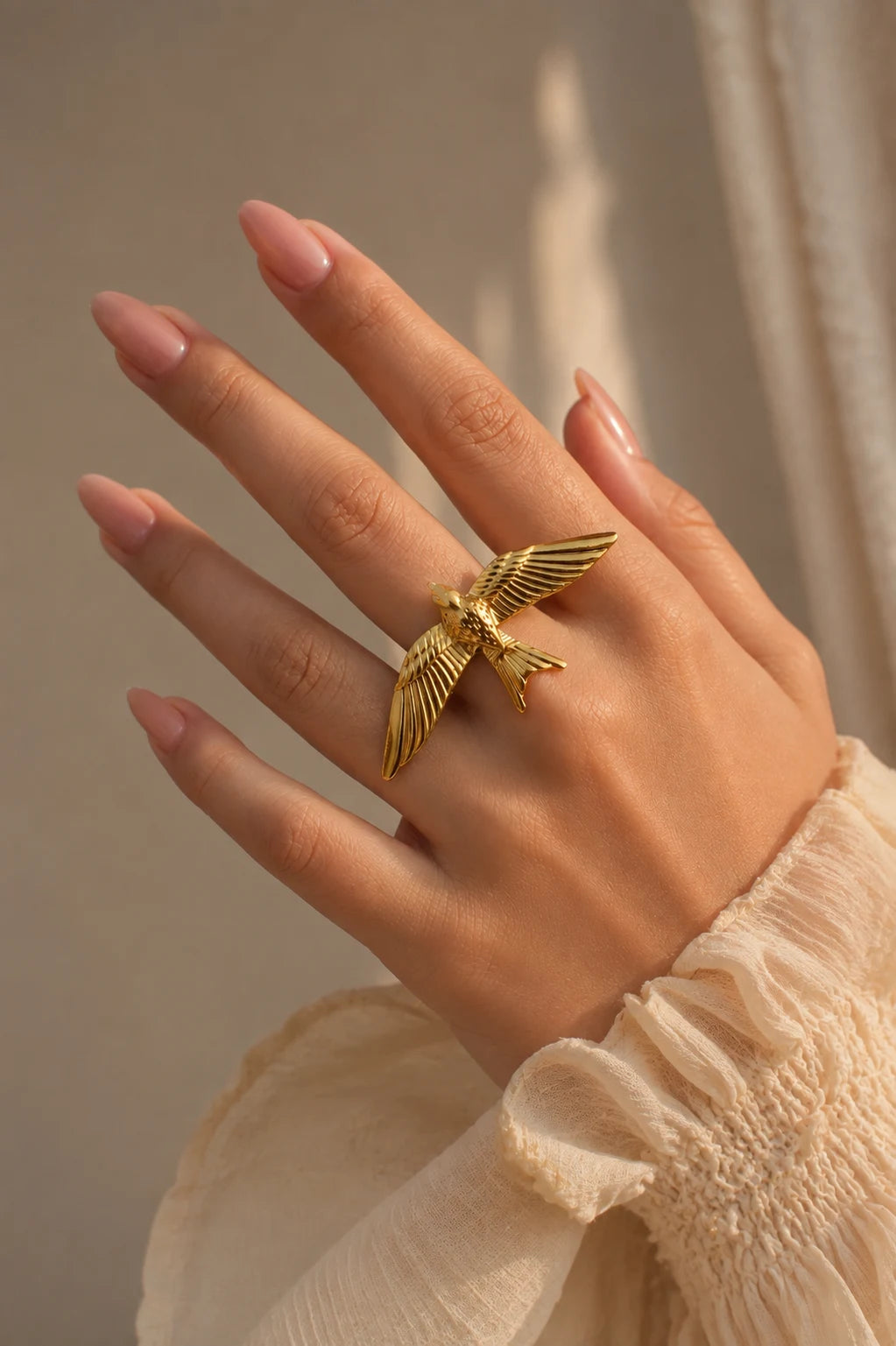 A gold bird ring worn on a hand with pink manicured nails, wearing a cream-colored ruffled blouse against a warm beige background.