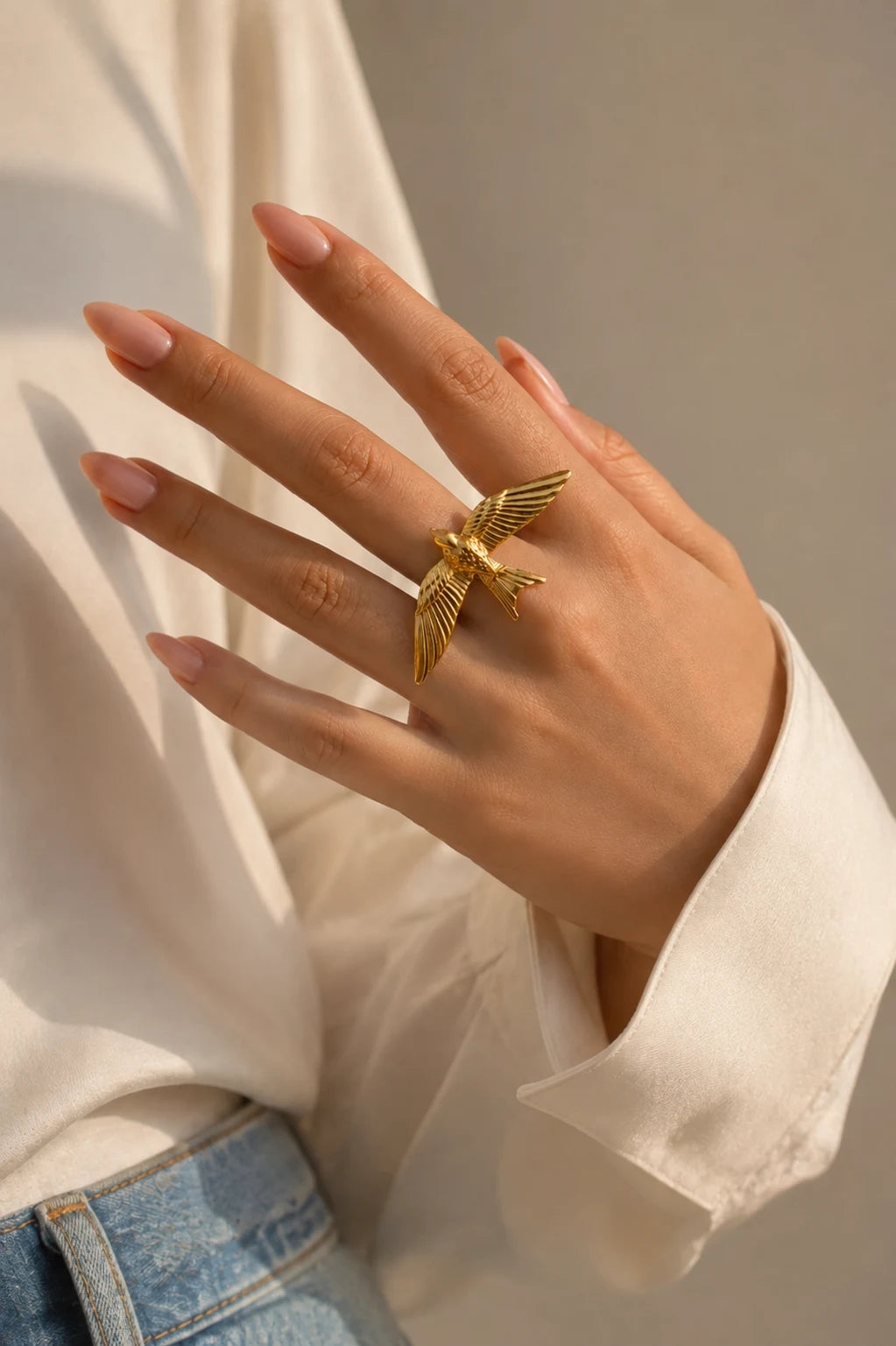 A gold bird ring worn on a person's hand, styled with a white blouse and blue jeans against a neutral background.