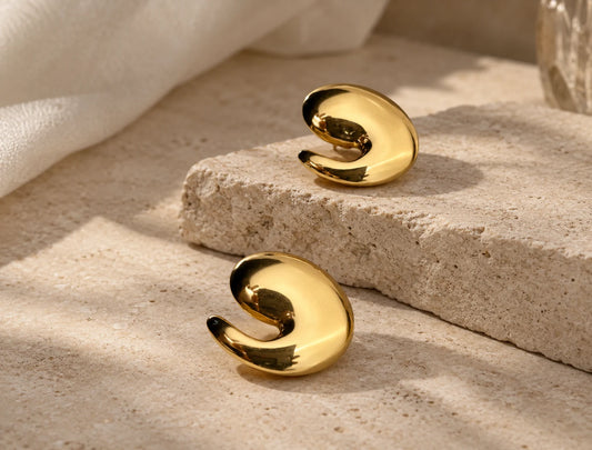 A pair of polished gold abstract spiral-shaped earrings displayed on a textured beige stone block with soft natural shadows.
